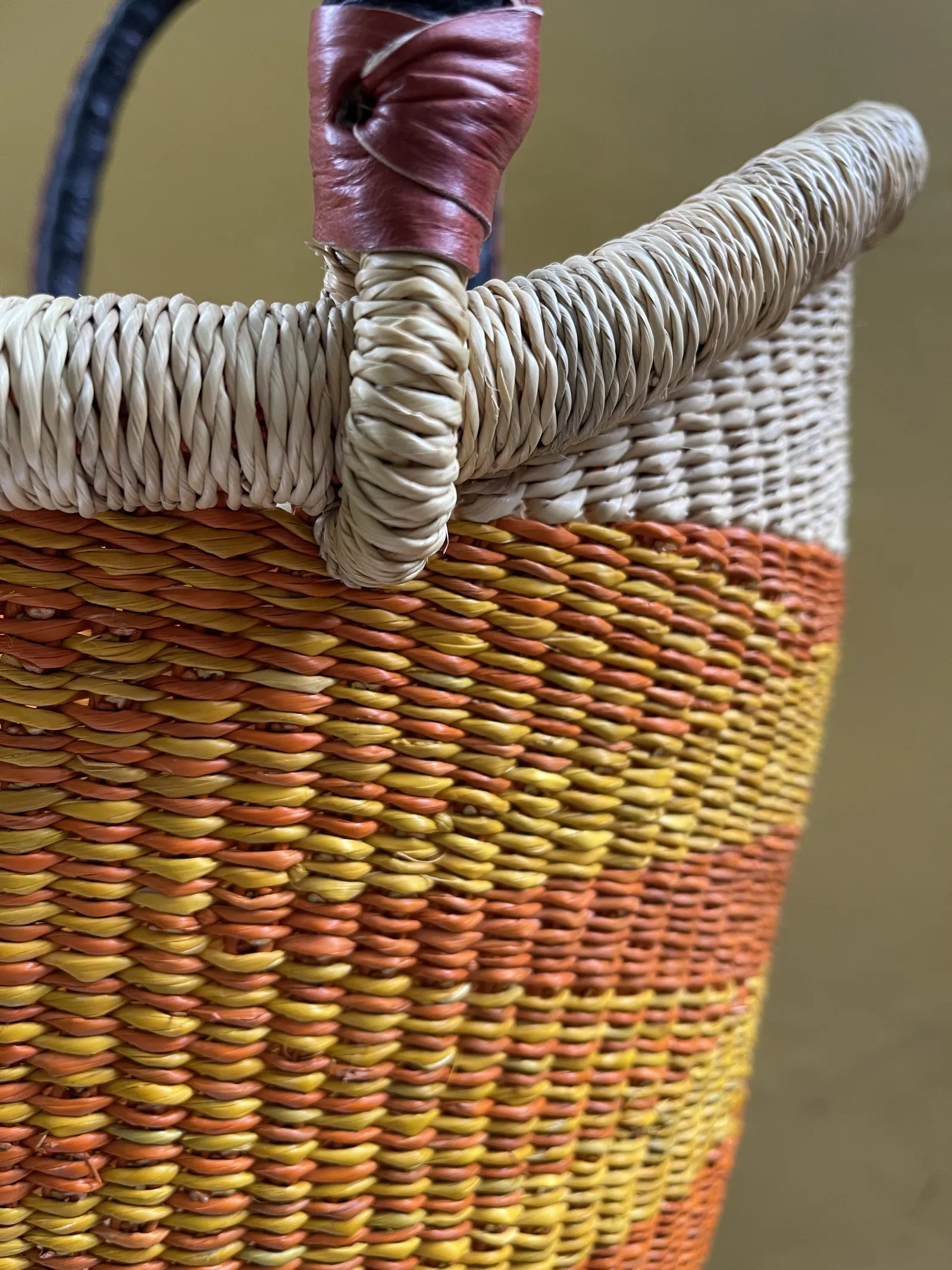 Close-up of a tightly wrapped basket rim and handle join above orange and yellow weaving.